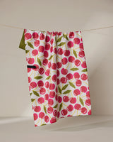 The Geometry Cherry Chic tea towel, featuring a red cherry and green leaf pattern, hangs on a thin string against a neutral background. Its quick-drying fabric combines function with style.