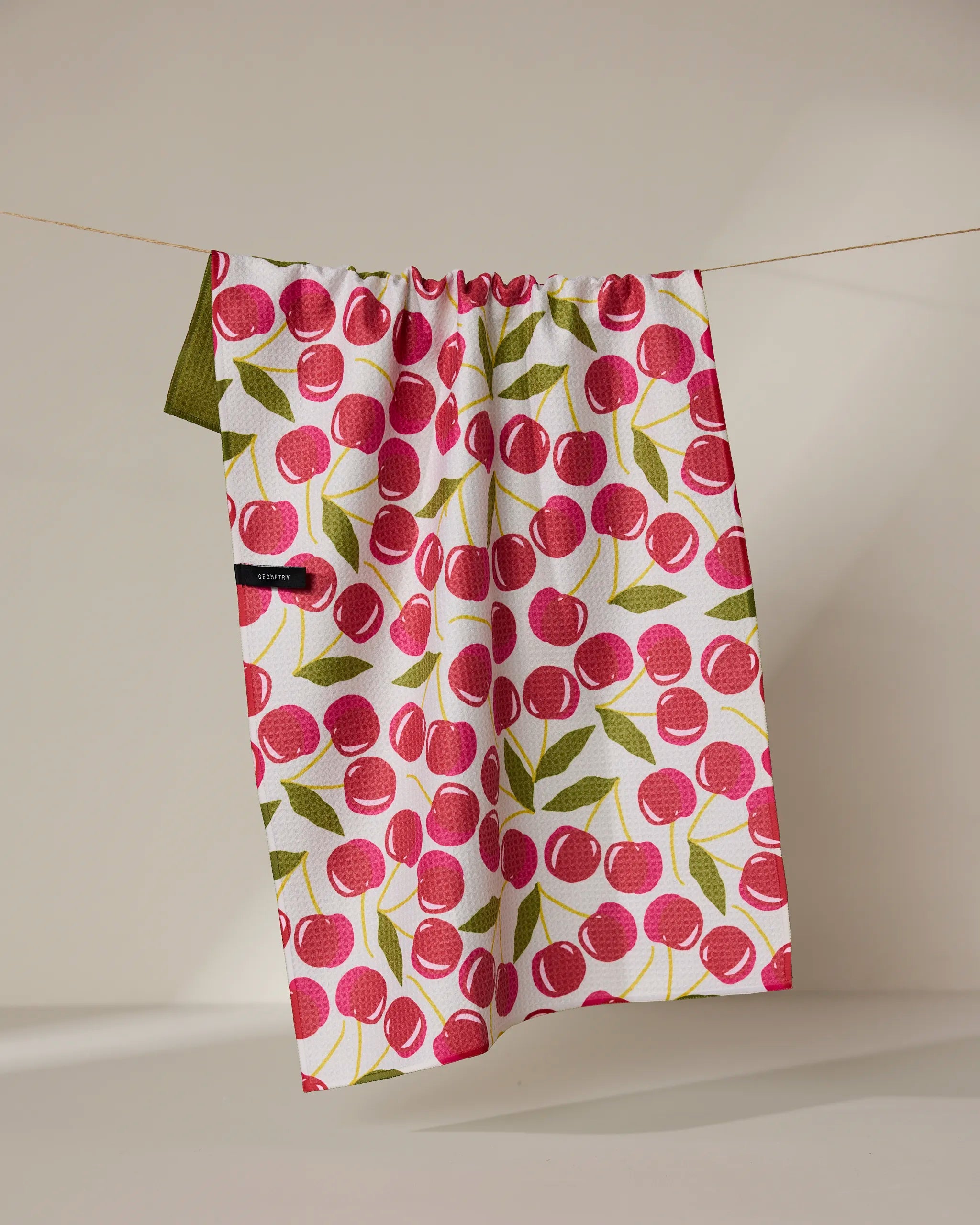The Geometry Cherry Chic tea towel, featuring a red cherry and green leaf pattern, hangs on a thin string against a neutral background. Its quick-drying fabric combines function with style.