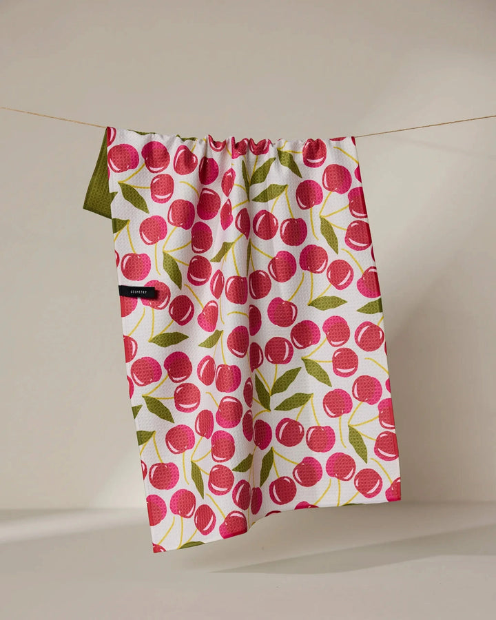 The Geometry Cherry Chic tea towel, featuring a red cherry and green leaf pattern, hangs on a thin string against a neutral background. Its quick-drying fabric combines function with style.