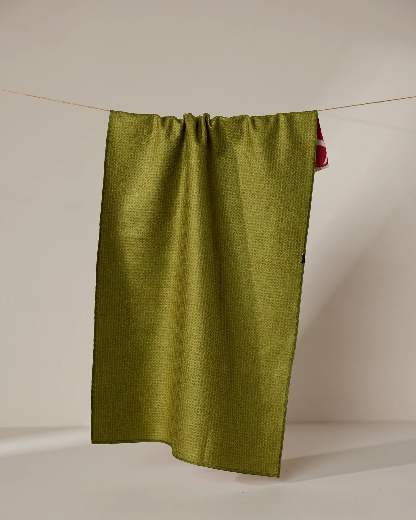 The Geometry Cherry Chic tea towel, featuring a green textured double-sided design, hangs neatly with a hint of red fabric peeking from the right, set against a plain light background.