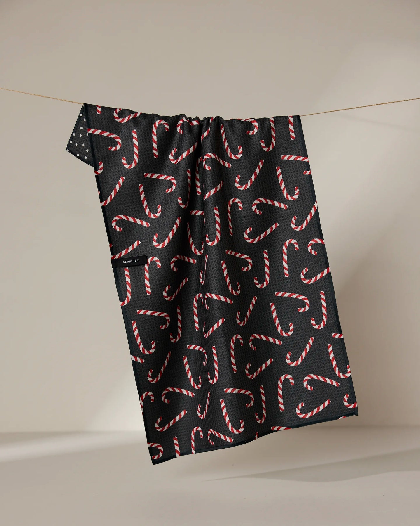 The Sweet Canes recycled kitchen towel by Geometry, featuring a red and white candy cane pattern on black, hangs on a string against a neutral background.