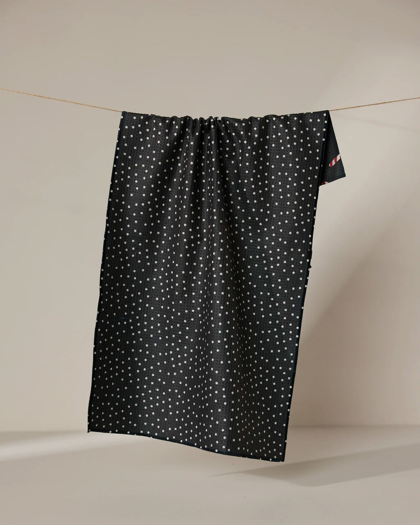 The Geometry Sweet Canes tea towel, featuring a dark background and small white dots, hangs on a clothesline against a plain beige backdrop. The quick-drying fabric drapes down in gentle folds.