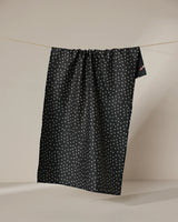 The Geometry Sweet Canes tea towel, featuring a dark background and small white dots, hangs on a clothesline against a plain beige backdrop. The quick-drying fabric drapes down in gentle folds.