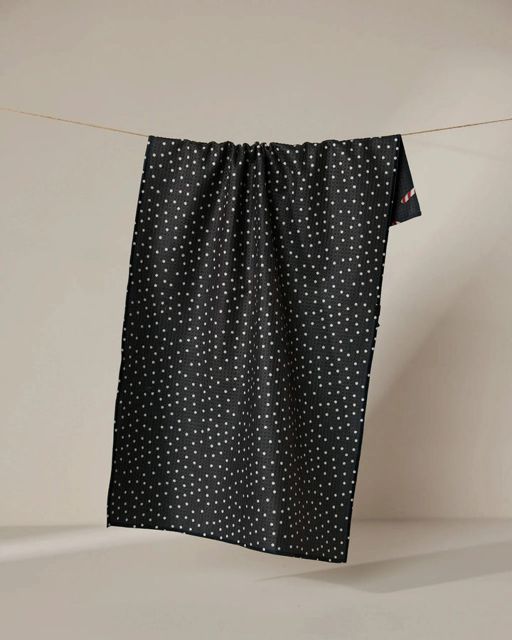 The Geometry Sweet Canes tea towel, featuring a dark background and small white dots, hangs on a clothesline against a plain beige backdrop. The quick-drying fabric drapes down in gentle folds.