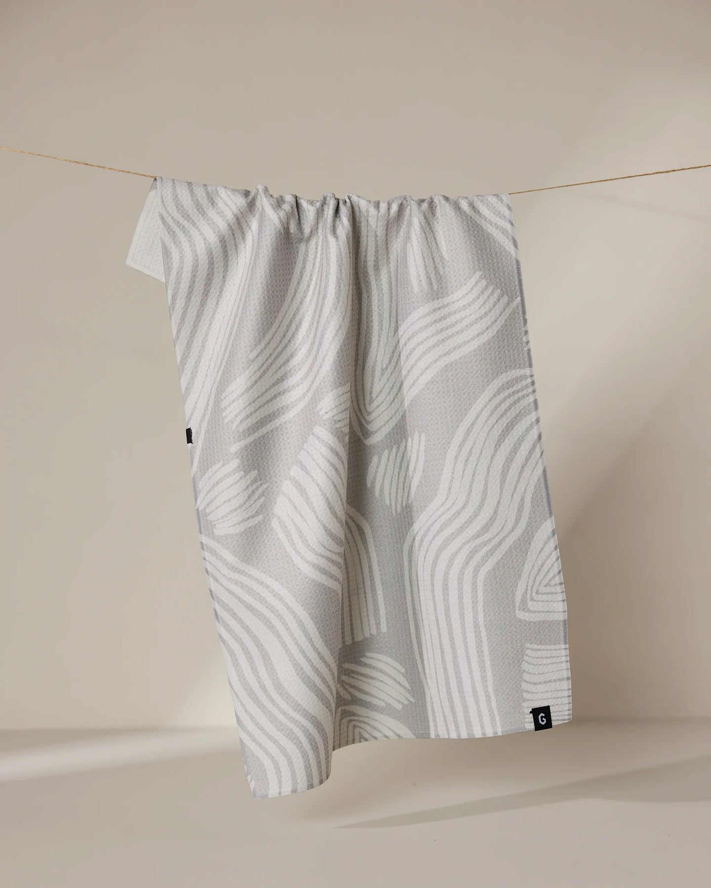 The Geometry Stone kitchen towel—light gray with abstract wavy white lines—hangs from a thin string against a plain beige background.