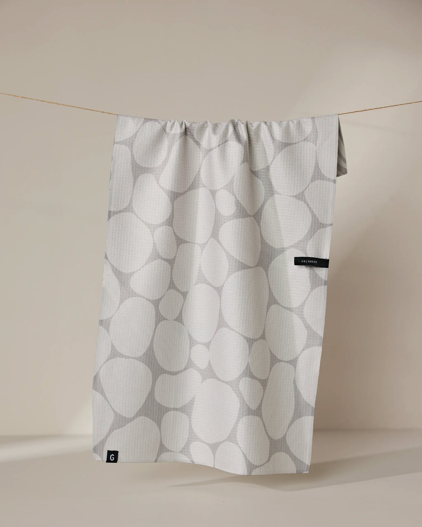 The Geometry Stone recycled kitchen towel, featuring a light gray pebble pattern and two black tags on one side, hangs from a thin string against a beige background.