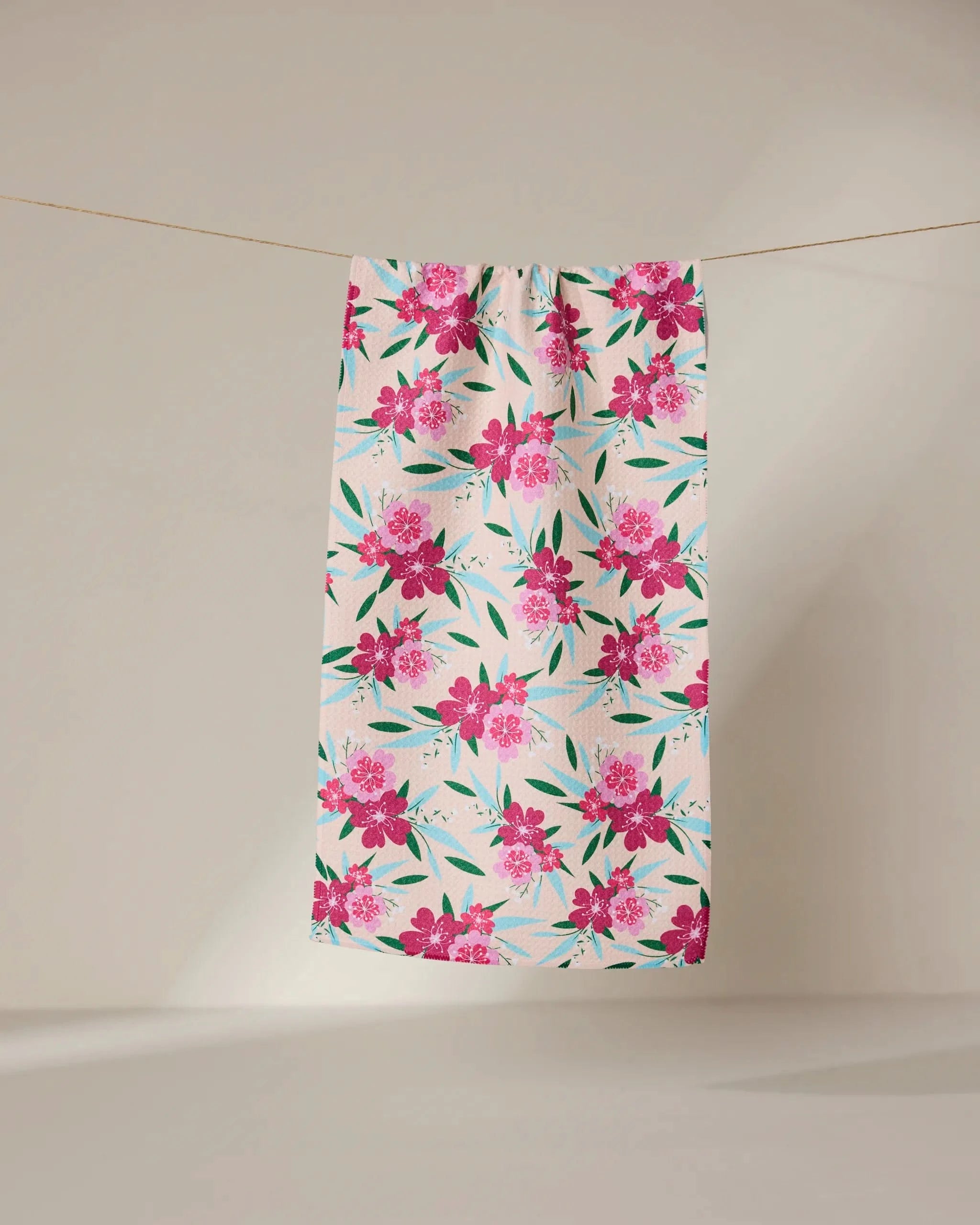 The GEOMETRY Sending Love bar towel features a white background with a bright pink floral pattern and green leaves. Made from ultra-absorbent fabric, it hangs on a clothesline against a neutral beige backdrop. Perfect for daily use.