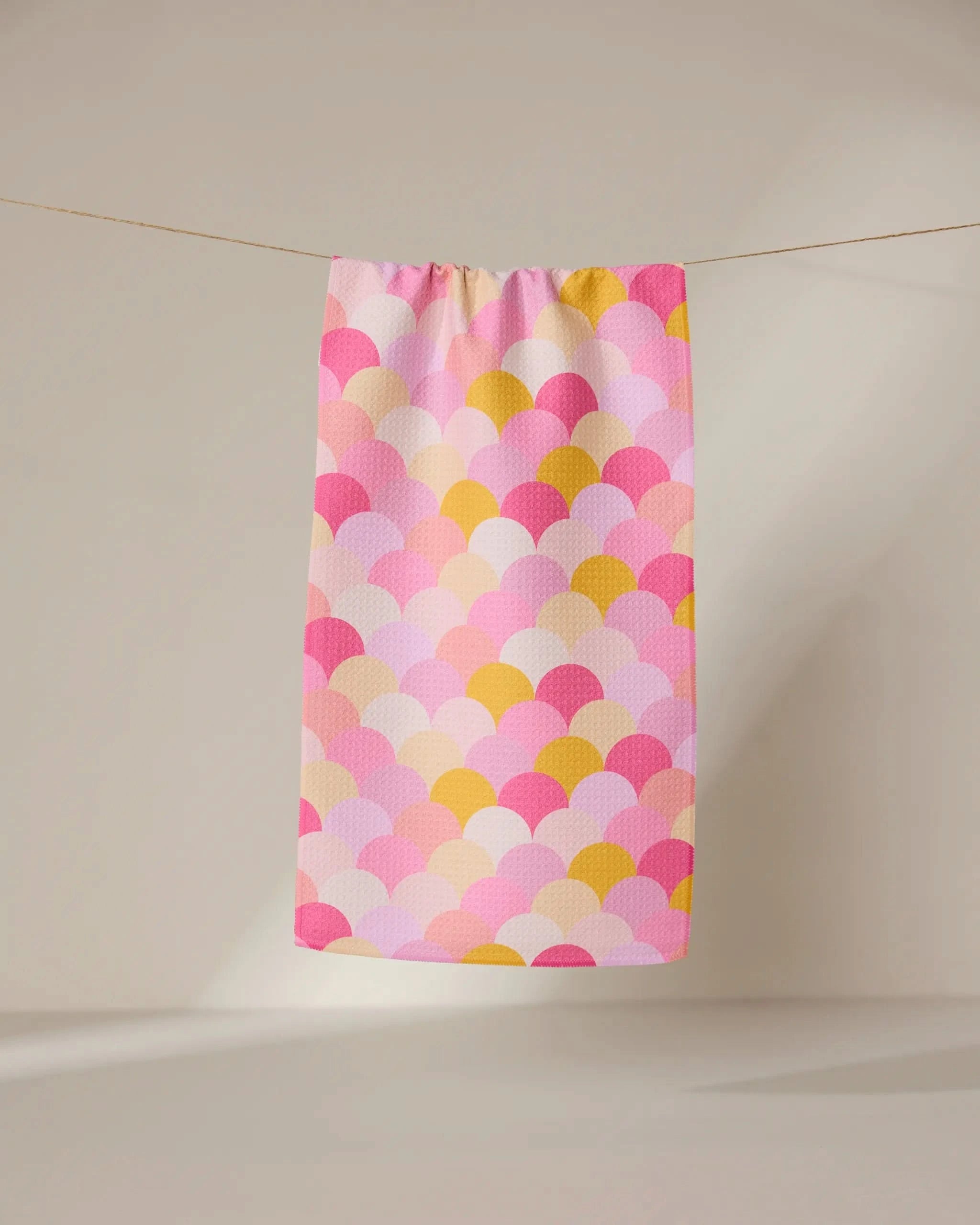 The GEOMETRY Inherit towel, featuring a scallop pattern in pink, yellow, and white, hangs on a clothesline against a neutral background.