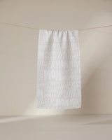 A white ultra-absorbent GEOMETRY Mom towel hangs on a line against a beige background, featuring “mom,” “mommy,” and “mother” repeatedly printed in large and small white and light gray lettering.