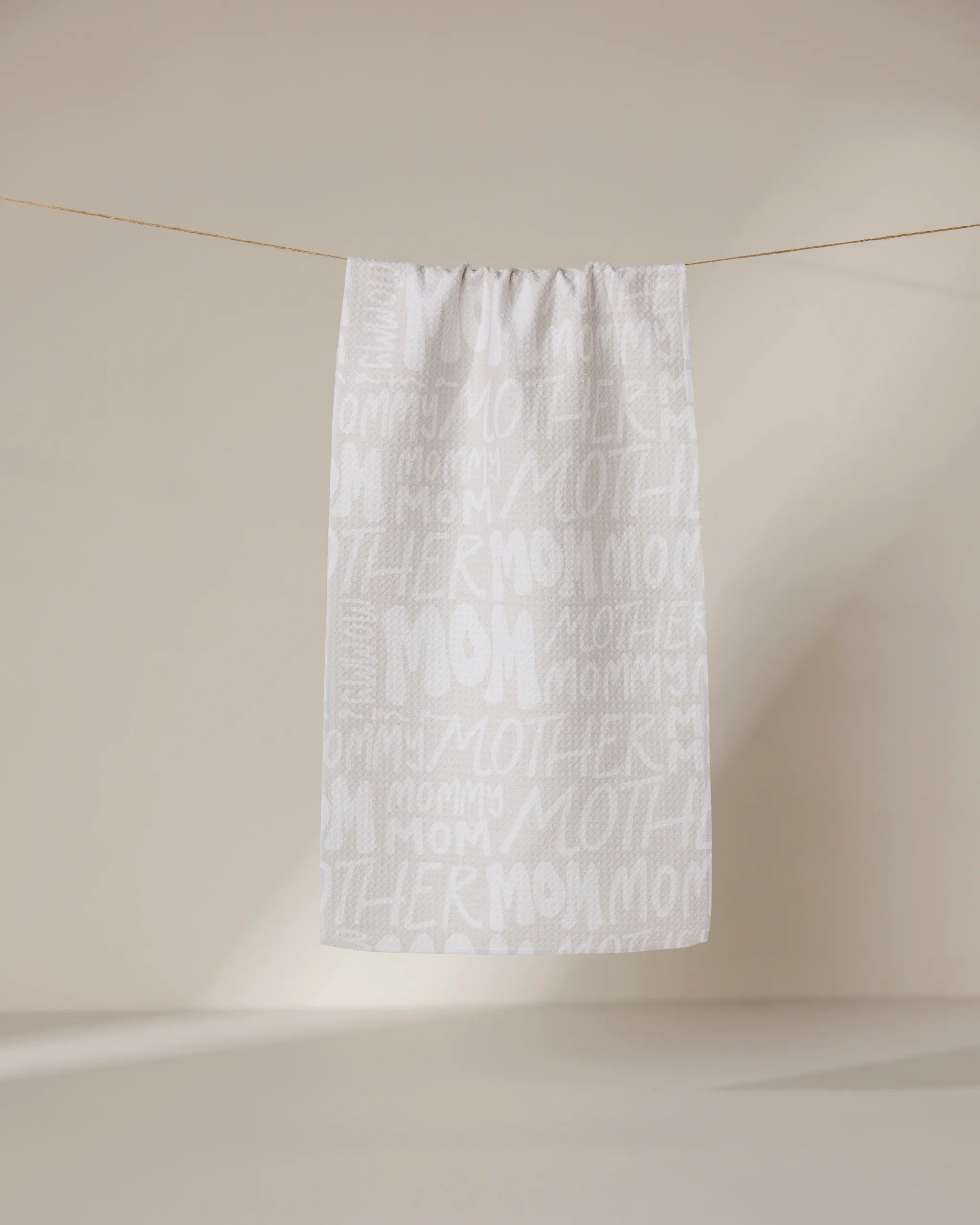 A white ultra-absorbent GEOMETRY Mom towel hangs on a line against a beige background, featuring “mom,” “mommy,” and “mother” repeatedly printed in large and small white and light gray lettering.