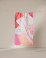 The GEOMETRY Memories towel, a rectangular ultra-absorbent piece featuring an abstract pink, orange, and white design with overlapping shapes and curved yellow lines, hangs on a clothesline against a neutral backdrop.