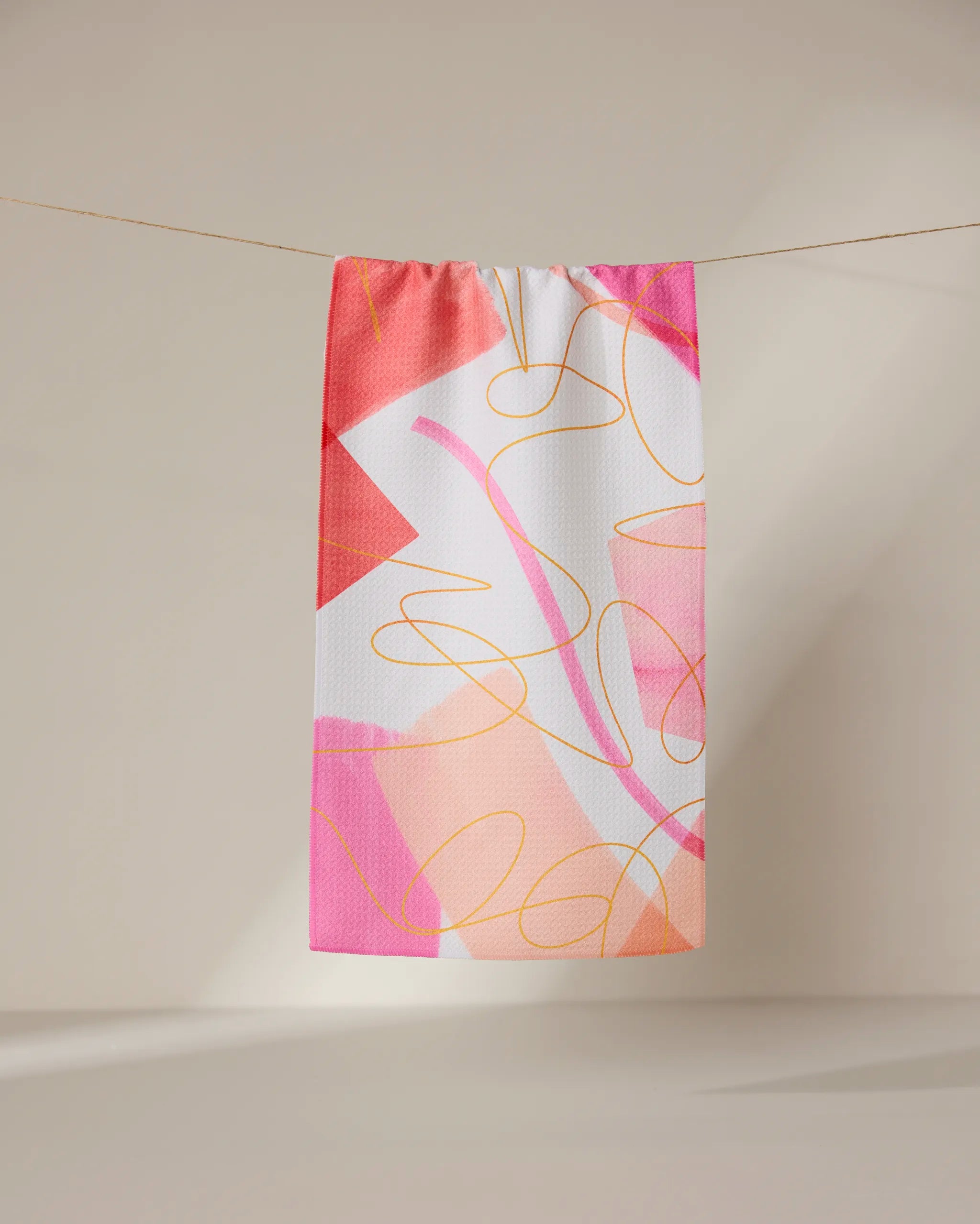 The GEOMETRY Memories towel, a rectangular ultra-absorbent piece featuring an abstract pink, orange, and white design with overlapping shapes and curved yellow lines, hangs on a clothesline against a neutral backdrop.