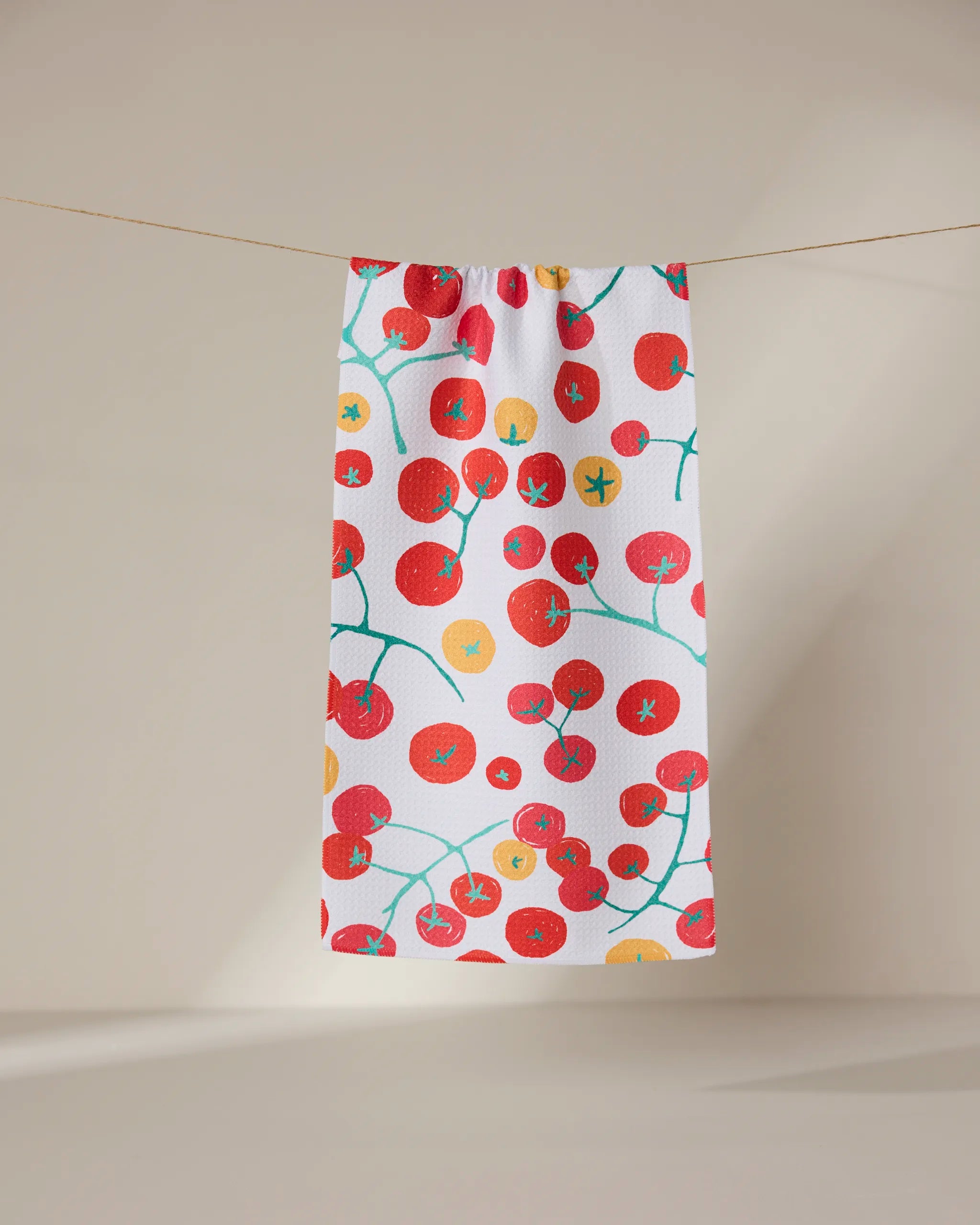 The GEOMETRY Ripe towel, ultra-absorbent and white with a vibrant tomato pattern of red and yellow tomatoes plus green vines, hangs on a string against a light beige background.