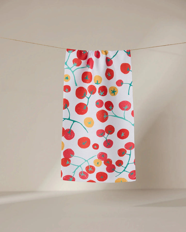 The GEOMETRY Ripe towel, ultra-absorbent and white with a vibrant tomato pattern of red and yellow tomatoes plus green vines, hangs on a string against a light beige background.