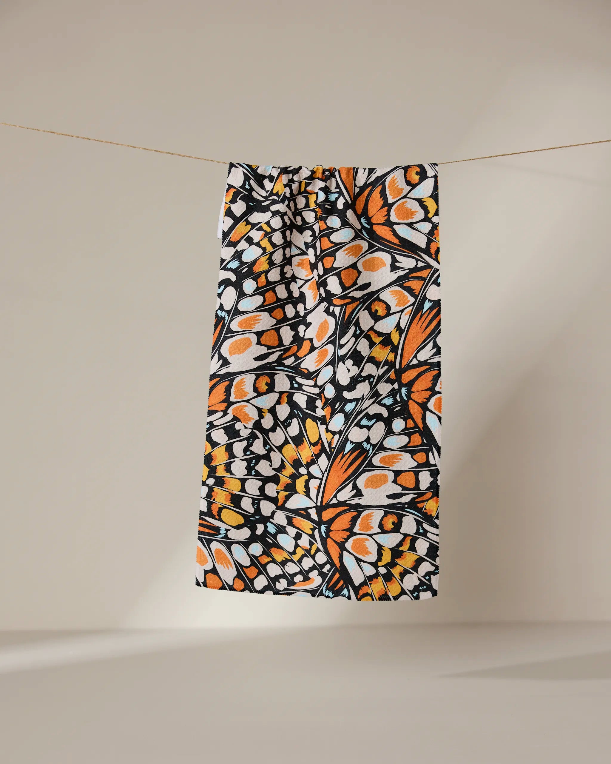 The GEOMETRY Wings in the Wind bar towel, featuring an orange, black, and white butterfly wing pattern, hangs on a thin line against a neutral background.