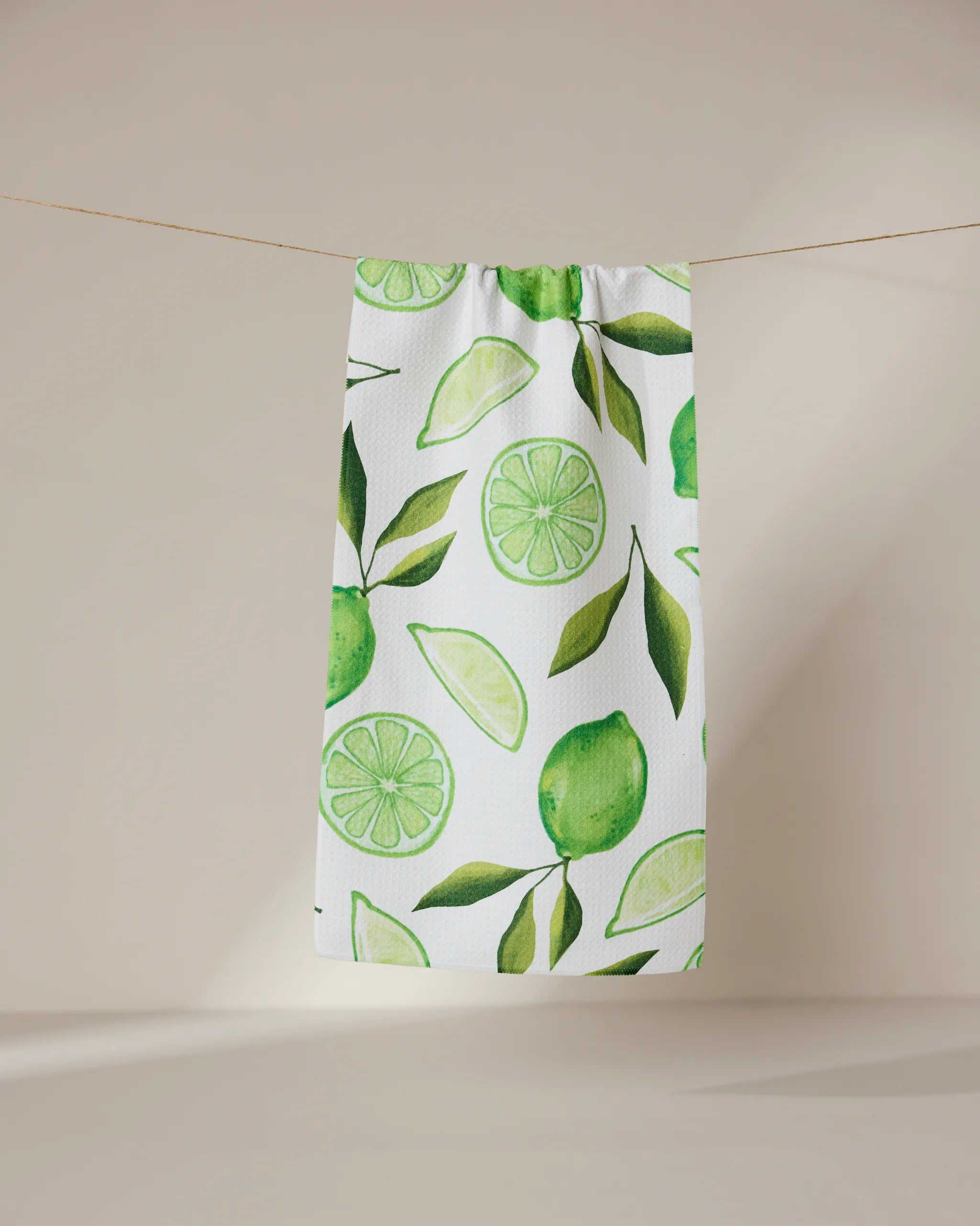 The GEOMETRY Lima kitchen towel, crafted from recycled materials and featuring green lime and leaf designs, hangs on a clothesline against a simple beige background.
