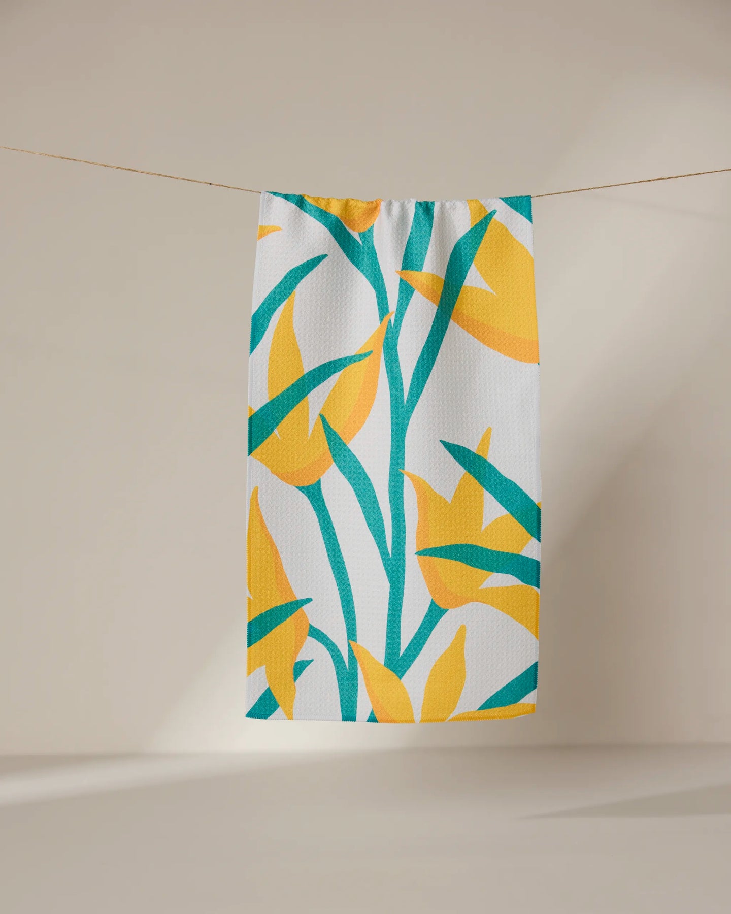 The GEOMETRY Growth towel, ultra-absorbent and white with vibrant yellow and green tropical leaf print, hangs on a clothesline against a neutral background.