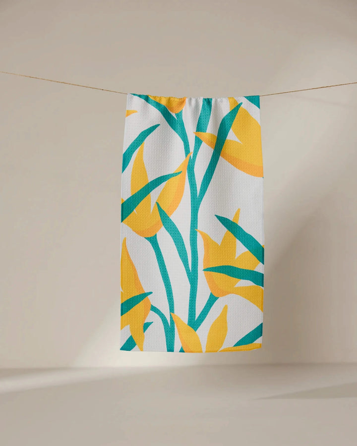 The GEOMETRY Growth towel, ultra-absorbent and white with vibrant yellow and green tropical leaf print, hangs on a clothesline against a neutral background.