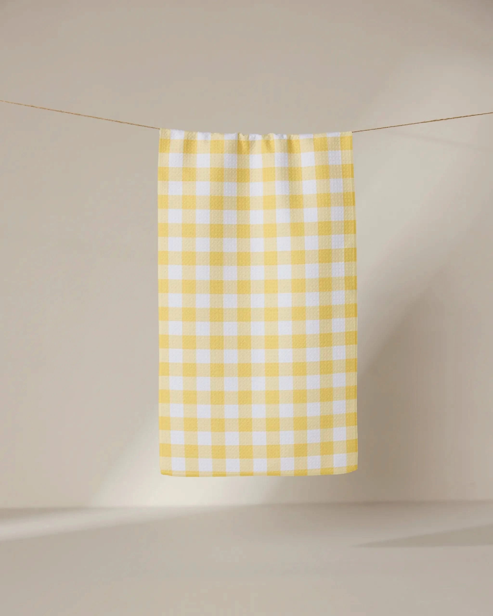 The GEOMETRY Lemon Gingham towel, featuring a yellow and white checkered design, hangs from a slender line against a neutral beige background, with soft lighting creating a minimalist atmosphere.
