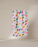 The GEOMETRY Downpour towel, ultra-absorbent and white with vibrant pink, orange, green, blue, and purple brushstroke patterns, hangs on a string against a softly lit plain background.