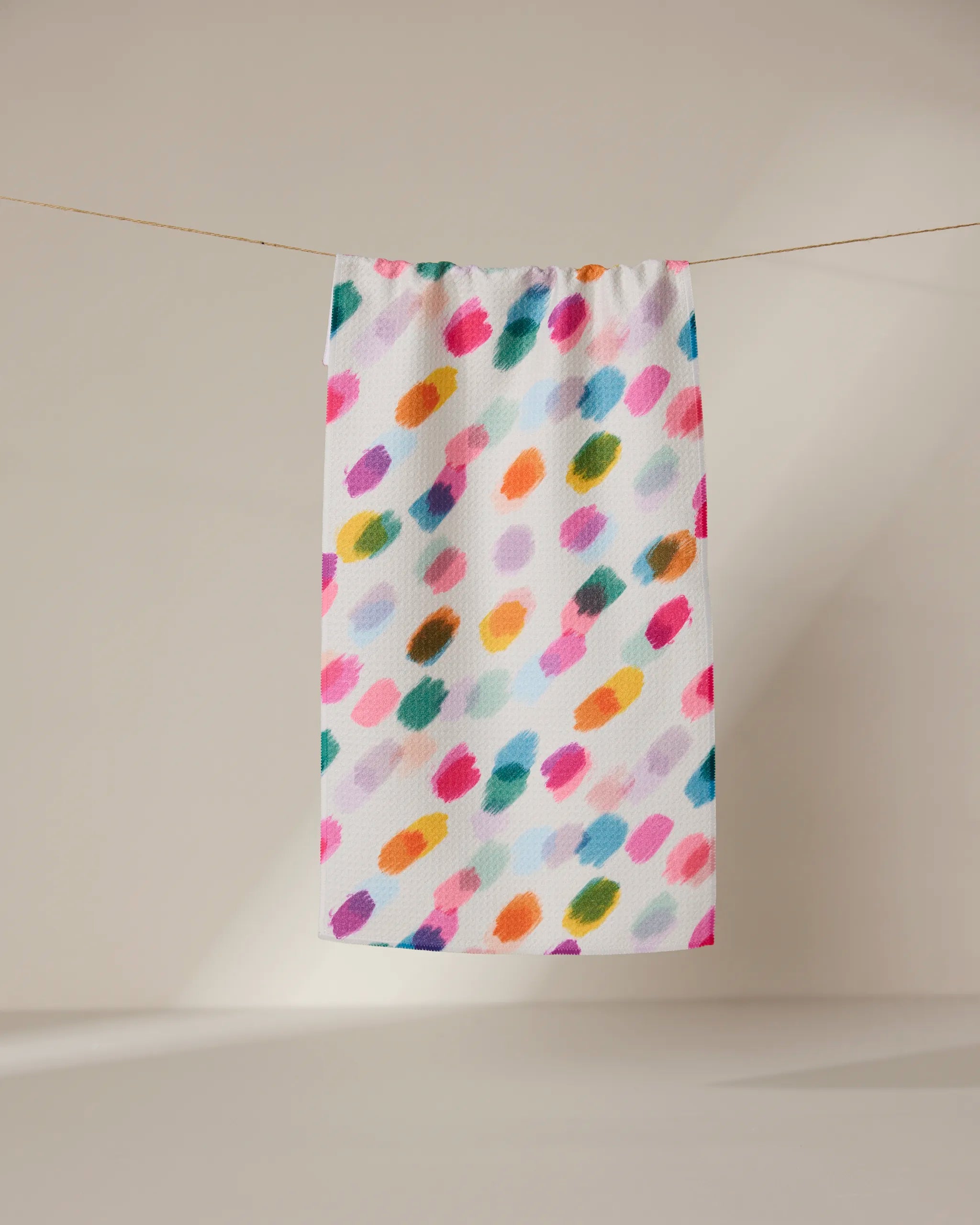 The GEOMETRY Downpour towel, ultra-absorbent and white with vibrant pink, orange, green, blue, and purple brushstroke patterns, hangs on a string against a softly lit plain background.