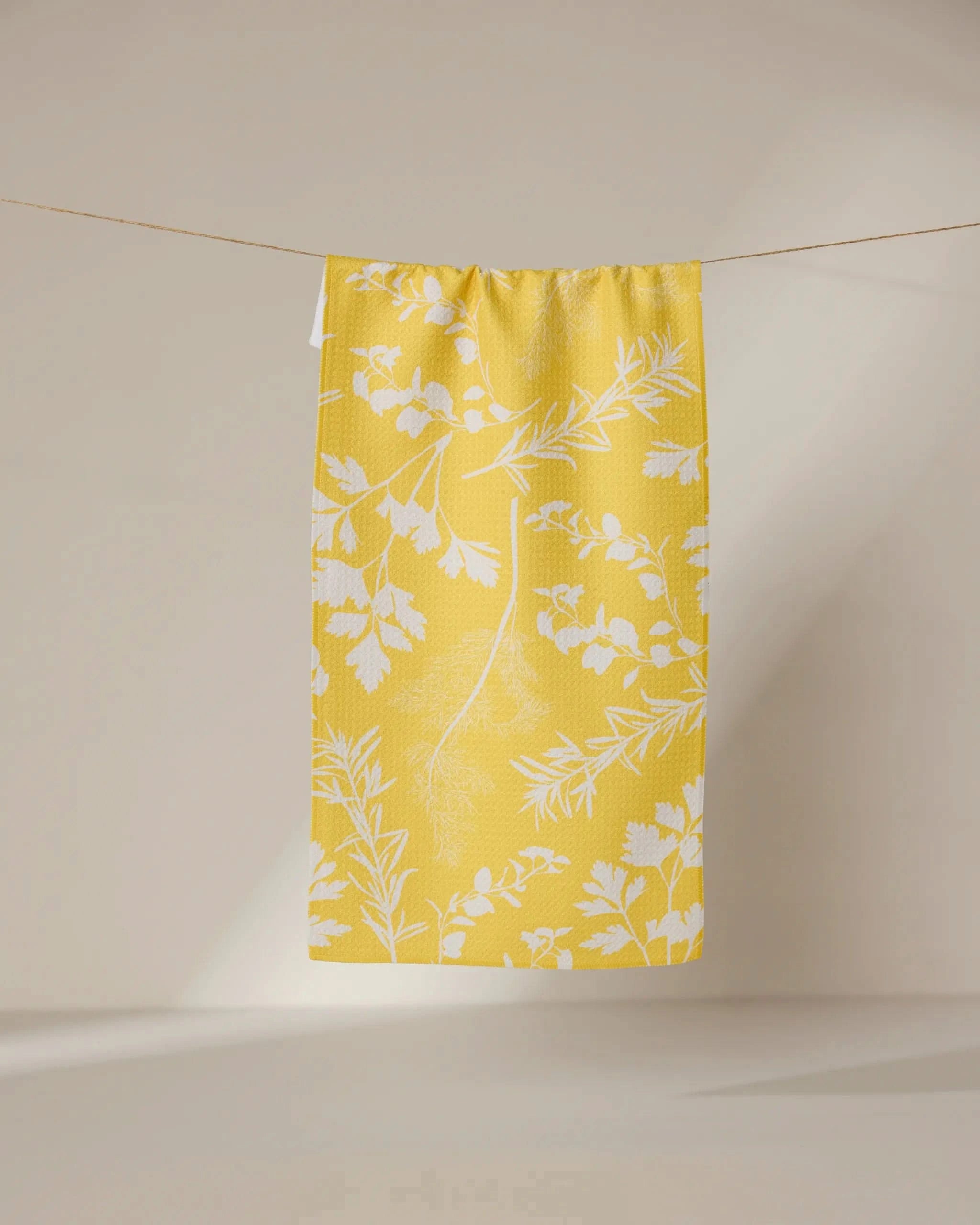 The GEOMETRY Garden of Gold bar towel, made from recycled materials, features a white botanical leaf and herb pattern on yellow and hangs on a string against a plain light background.