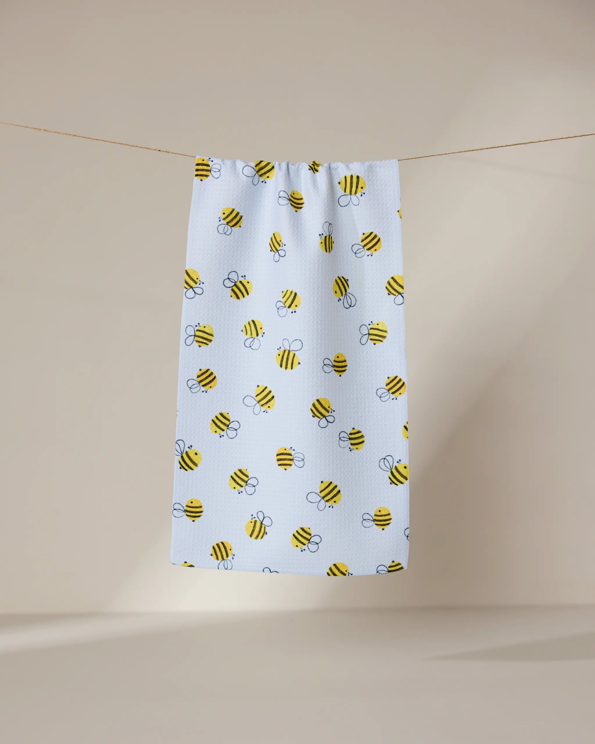 The GEOMETRY Buzzing towel, ultra-absorbent and white with yellow and black cartoon bee patterns, hangs on a clothesline against a plain beige background.