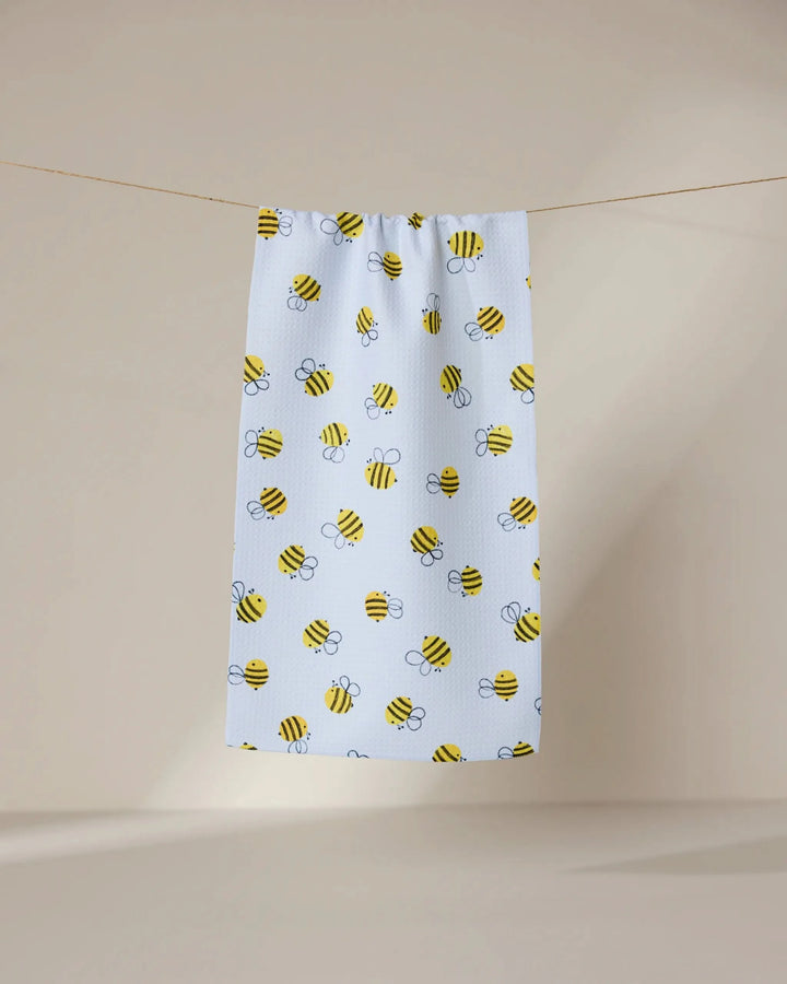 The GEOMETRY Buzzing towel, ultra-absorbent and white with yellow and black cartoon bee patterns, hangs on a clothesline against a plain beige background.