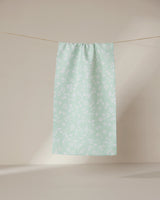 The GEOMETRY Dandy towel, in light green with a subtle white floral pattern, hangs on a clothesline against a neutral beige background.