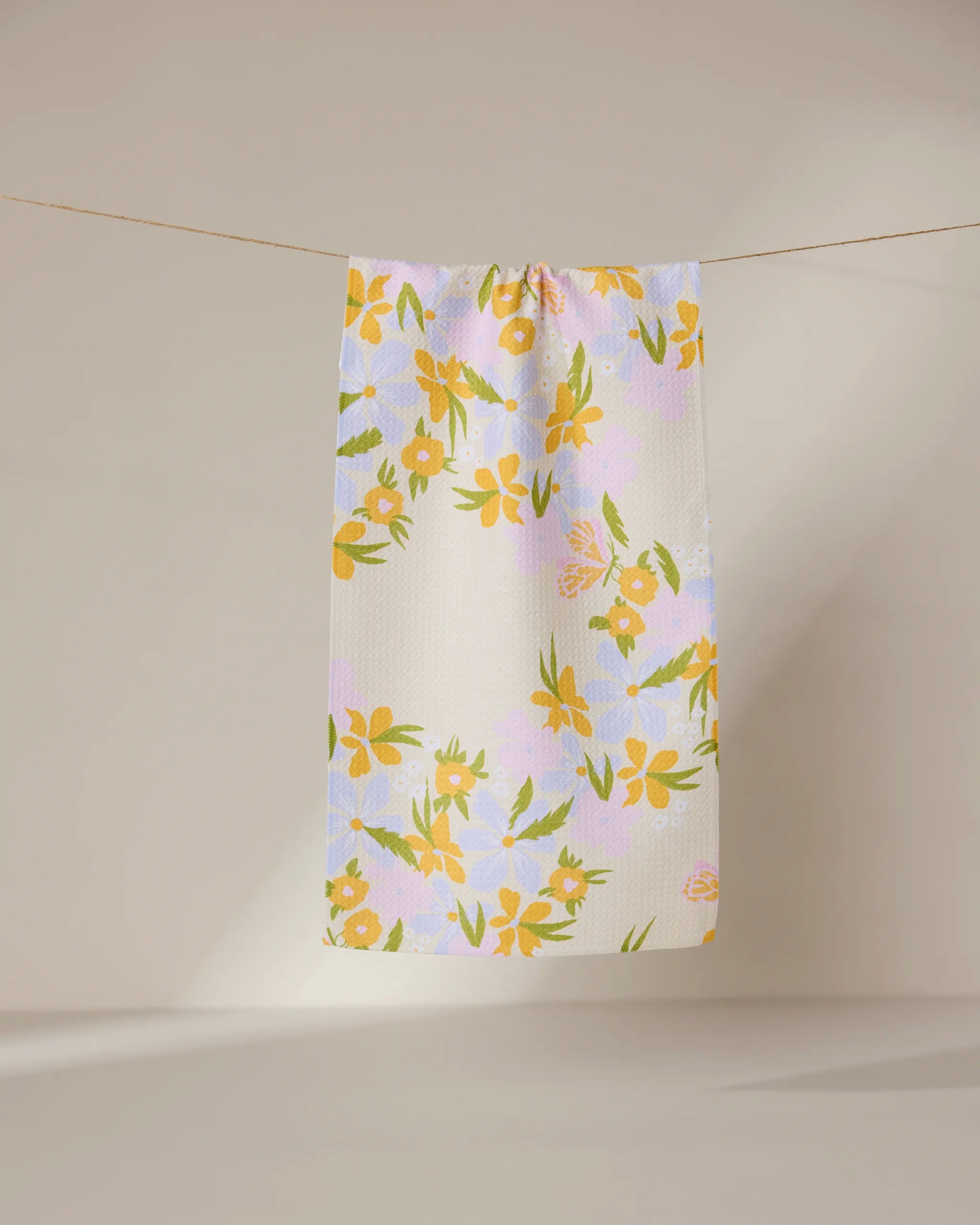 The GEOMETRY Abloom ultra-absorbent white bar towel, featuring a yellow and green floral pattern, hangs from a clothesline against a neutral background.
