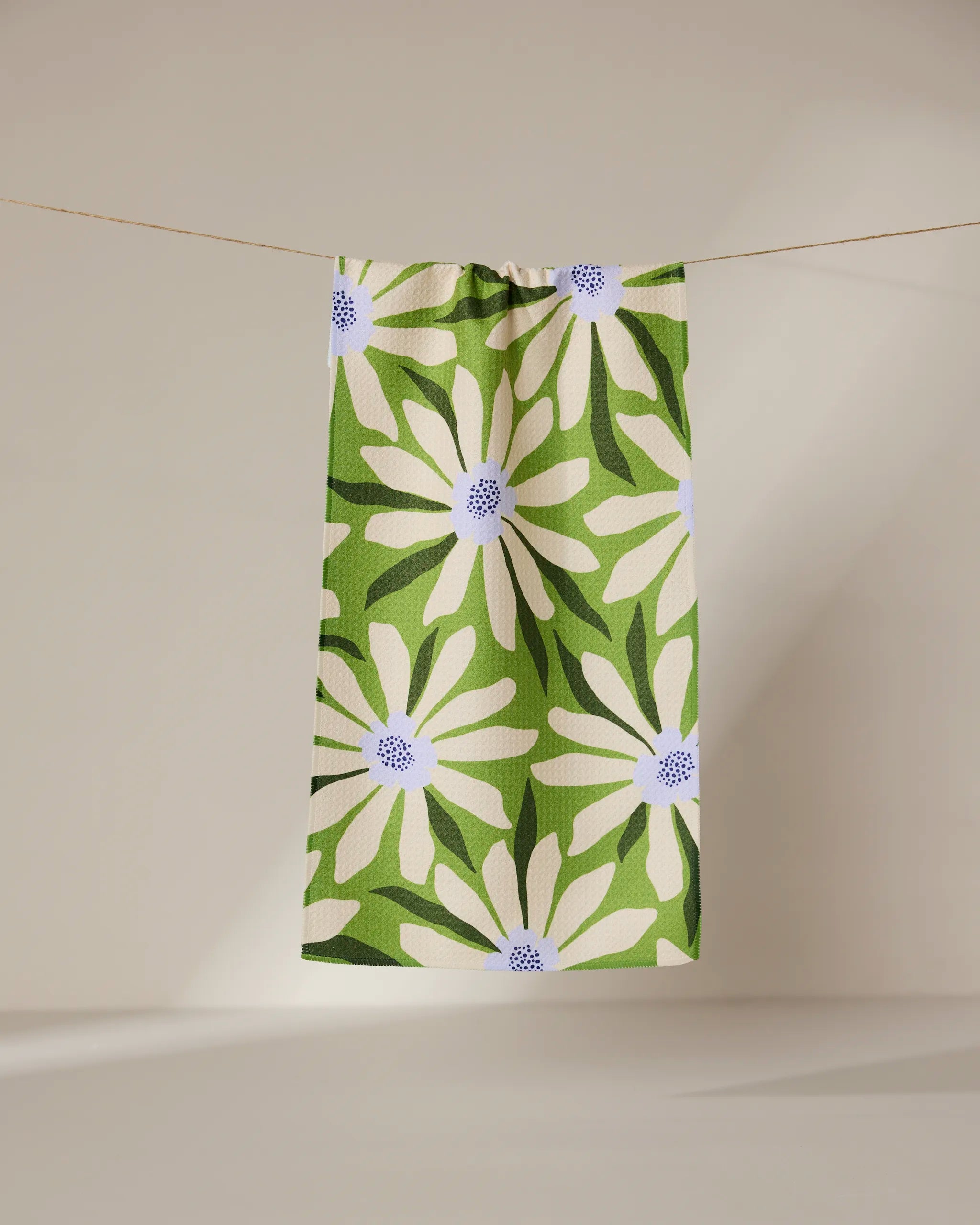 The GEOMETRY Bliss and Bloom green recycled kitchen towel, featuring a striking white daisy with a blue center, hangs on a clothesline against a neutral background.