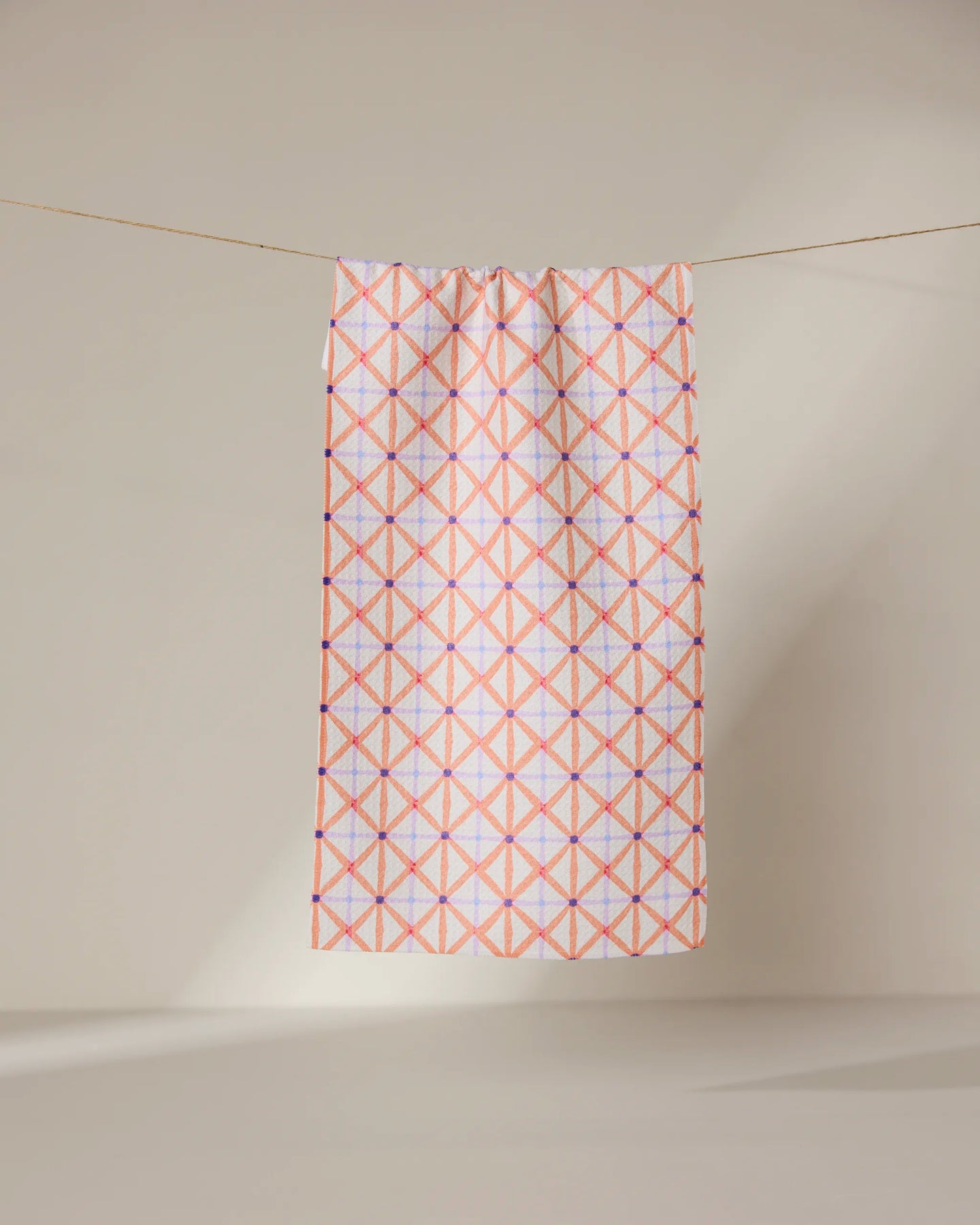The GEOMETRY Anew towel features ultra-absorbent fabric and a geometric pattern of intersecting pink and orange lines forming diamonds, shown hanging on a line against a neutral, light background.