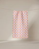 The GEOMETRY Anew towel features ultra-absorbent fabric and a geometric pattern of intersecting pink and orange lines forming diamonds, shown hanging on a line against a neutral, light background.