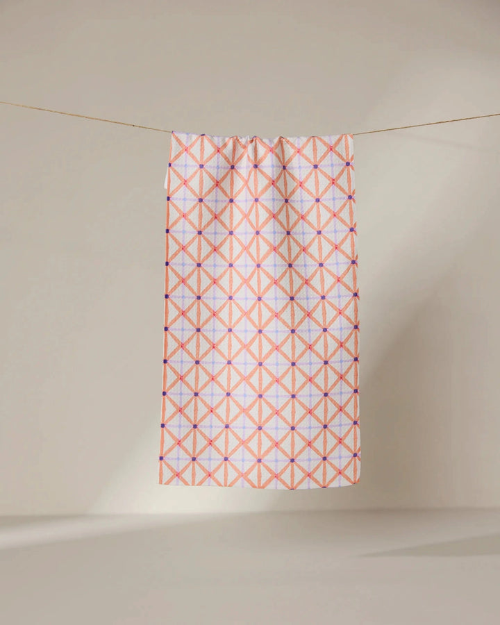 The GEOMETRY Anew towel features ultra-absorbent fabric and a geometric pattern of intersecting pink and orange lines forming diamonds, shown hanging on a line against a neutral, light background.