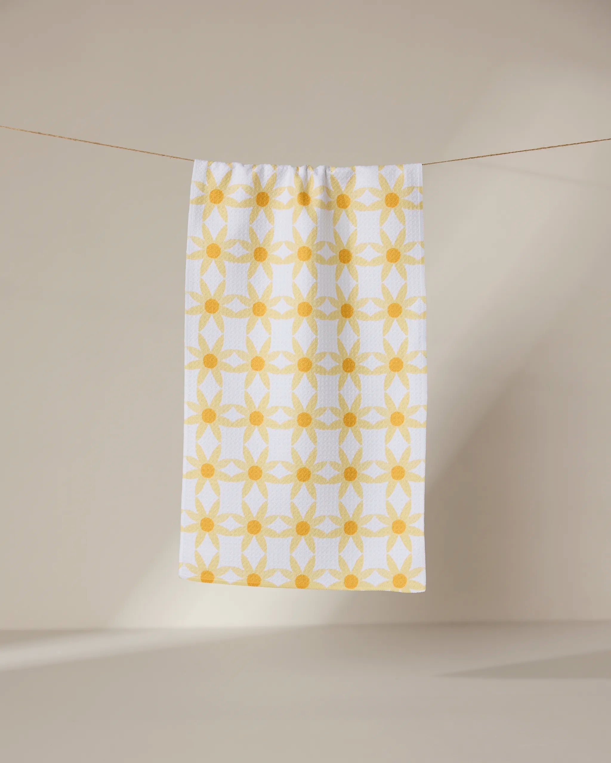 The GEOMETRY Daisies towel, featuring a yellow geometric floral pattern, hangs on a thin clothesline against a neutral beige background.