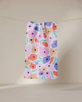 The Blossoming Love towel by GEOMETRY, featuring a bold abstract floral design in purple, orange, pink, and blue hues, hangs from a line against a neutral backdrop.
