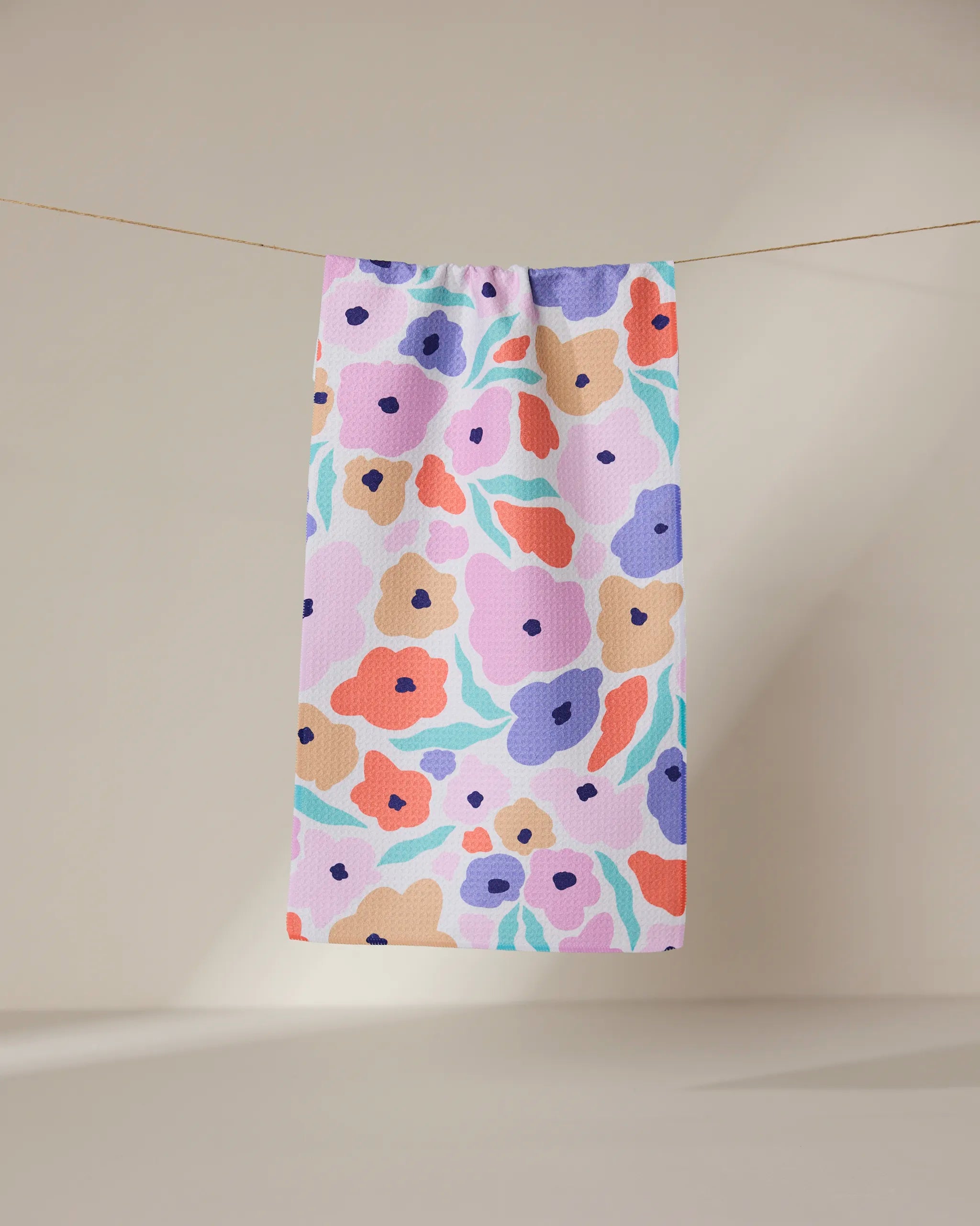 The Blossoming Love towel by GEOMETRY, featuring a bold abstract floral design in purple, orange, pink, and blue hues, hangs from a line against a neutral backdrop.