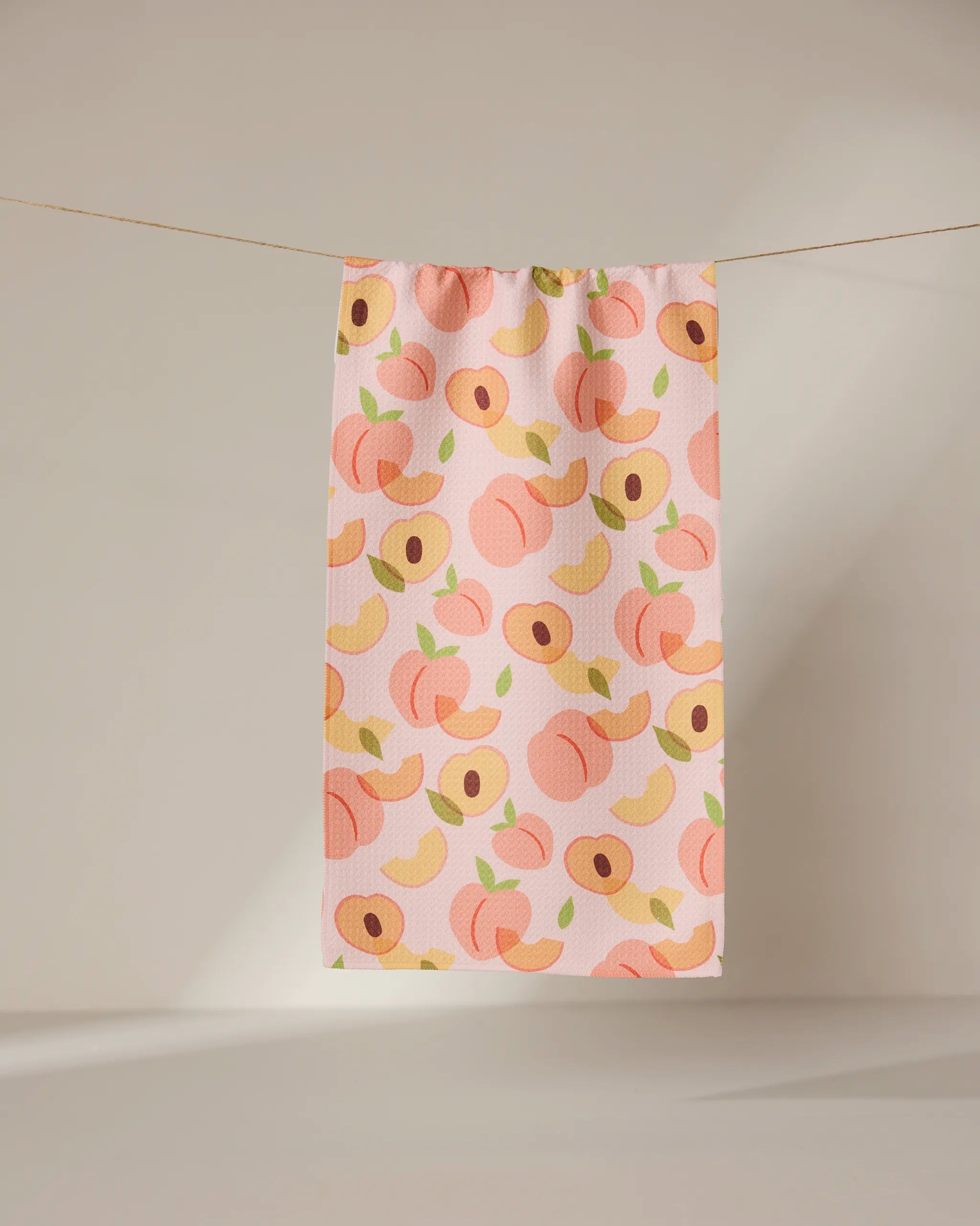 The GEOMETRY Peaches towel, in light pink and adorned with whole and sliced peaches in pink, yellow, and green, hangs on a clothesline against a neutral backdrop.