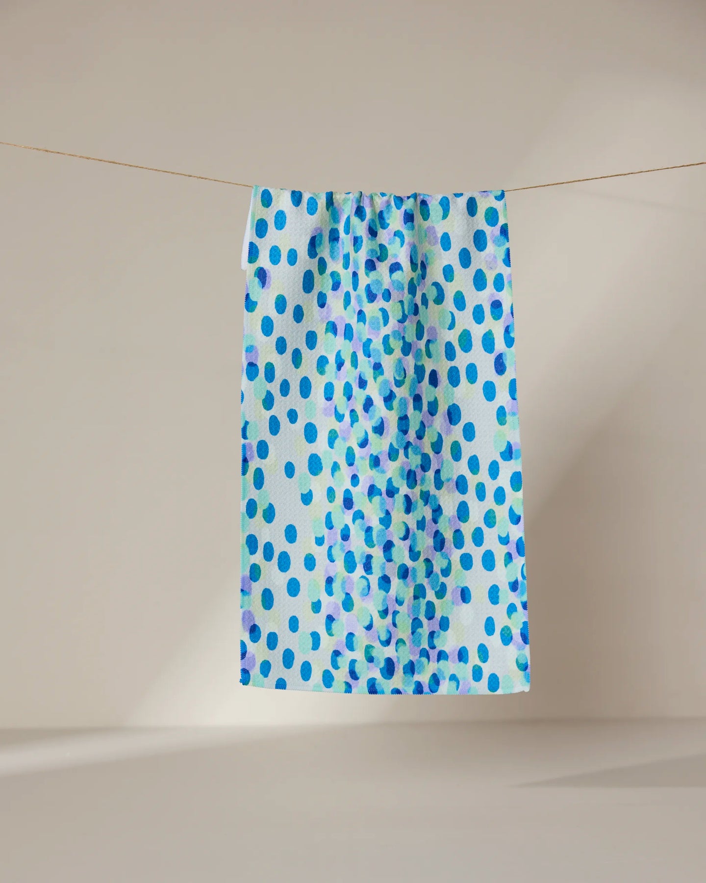 The GEOMETRY Spotty Summer towel, featuring a blue polka dot design and ultra-absorbent rectangular shape, hangs on a clothesline against a softly lit neutral background.