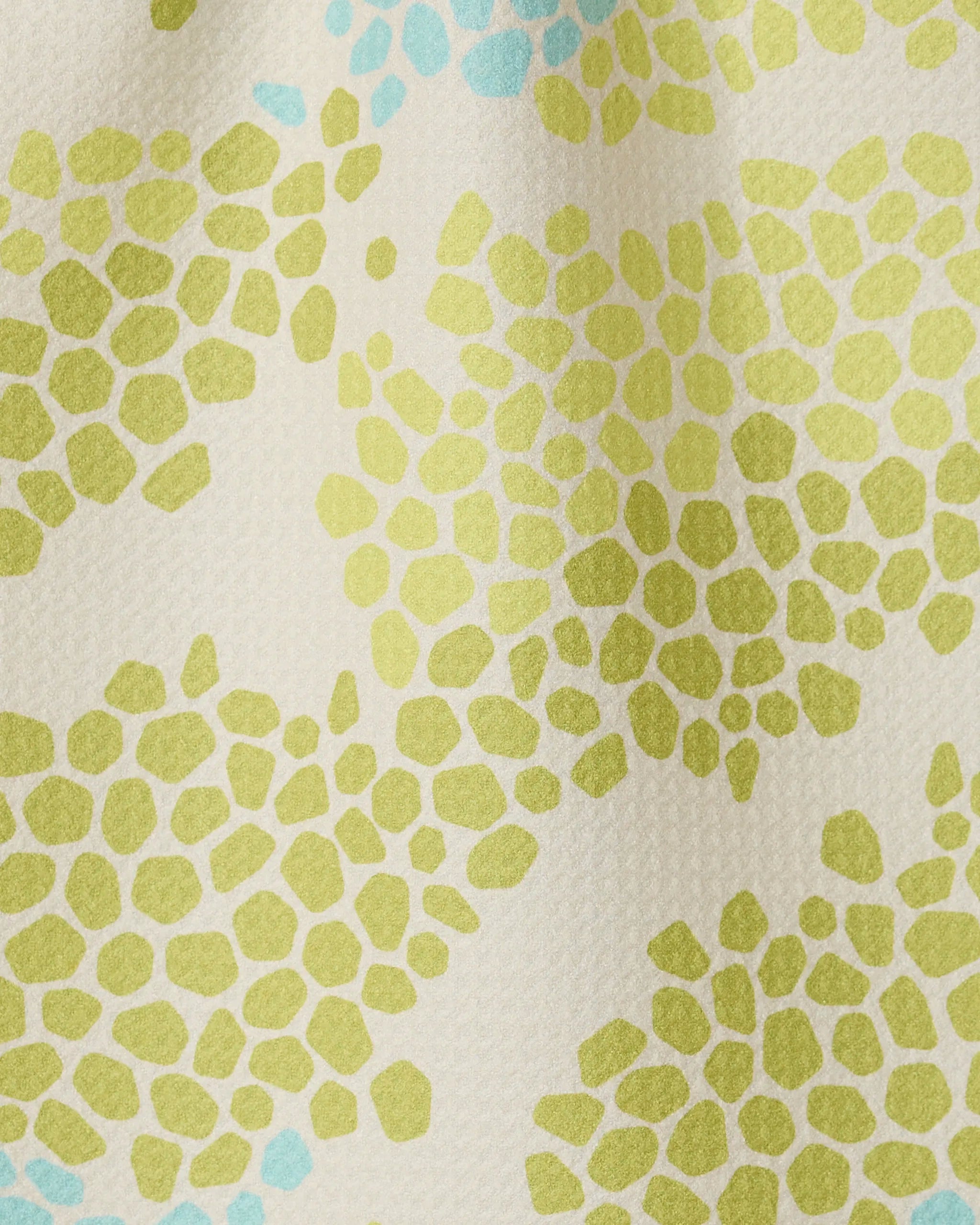 A close-up of GEOMETRY’s Geo Shell fabric shows clustered green and light blue irregular dots on a cream background, crafted from recycled materials and resembling abstract floral or pebble shapes.