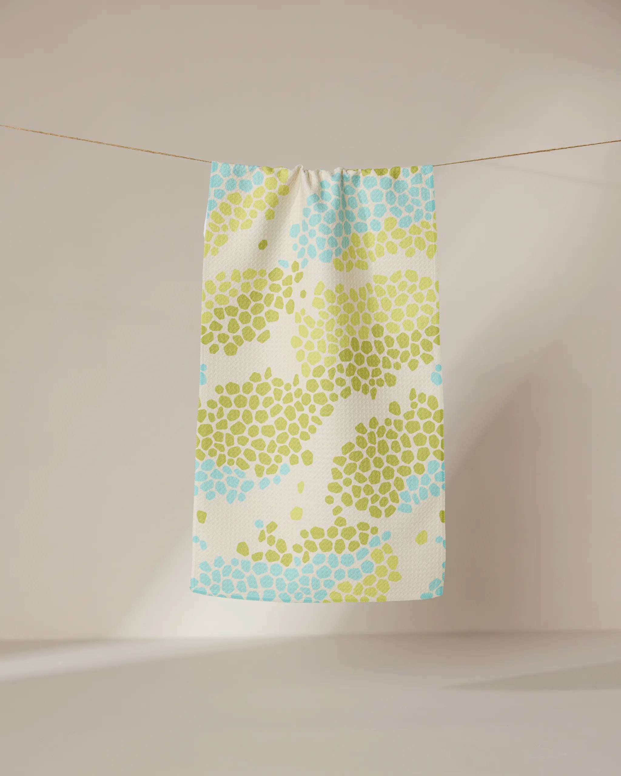 The GEOMETRY Geo Shell towel, featuring a patterned ultra-absorbent design, hangs on a clothesline with sunlight casting soft shadows on the neutral wall and floor in the background.