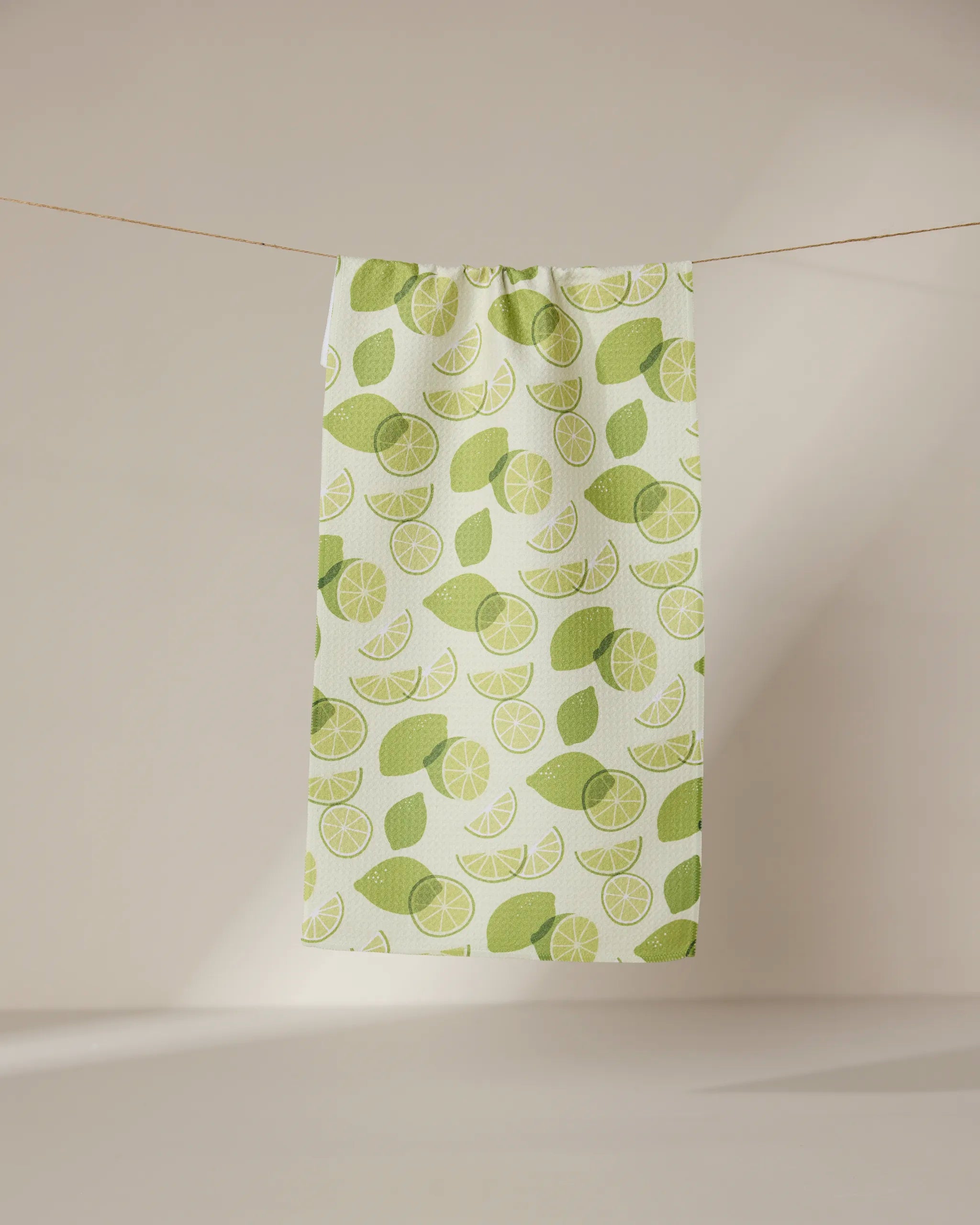 A white ultra-absorbent GEOMETRY towel named Limes, featuring a green lime and leaf pattern, hangs from a clothesline against a softly lit, plain background.
