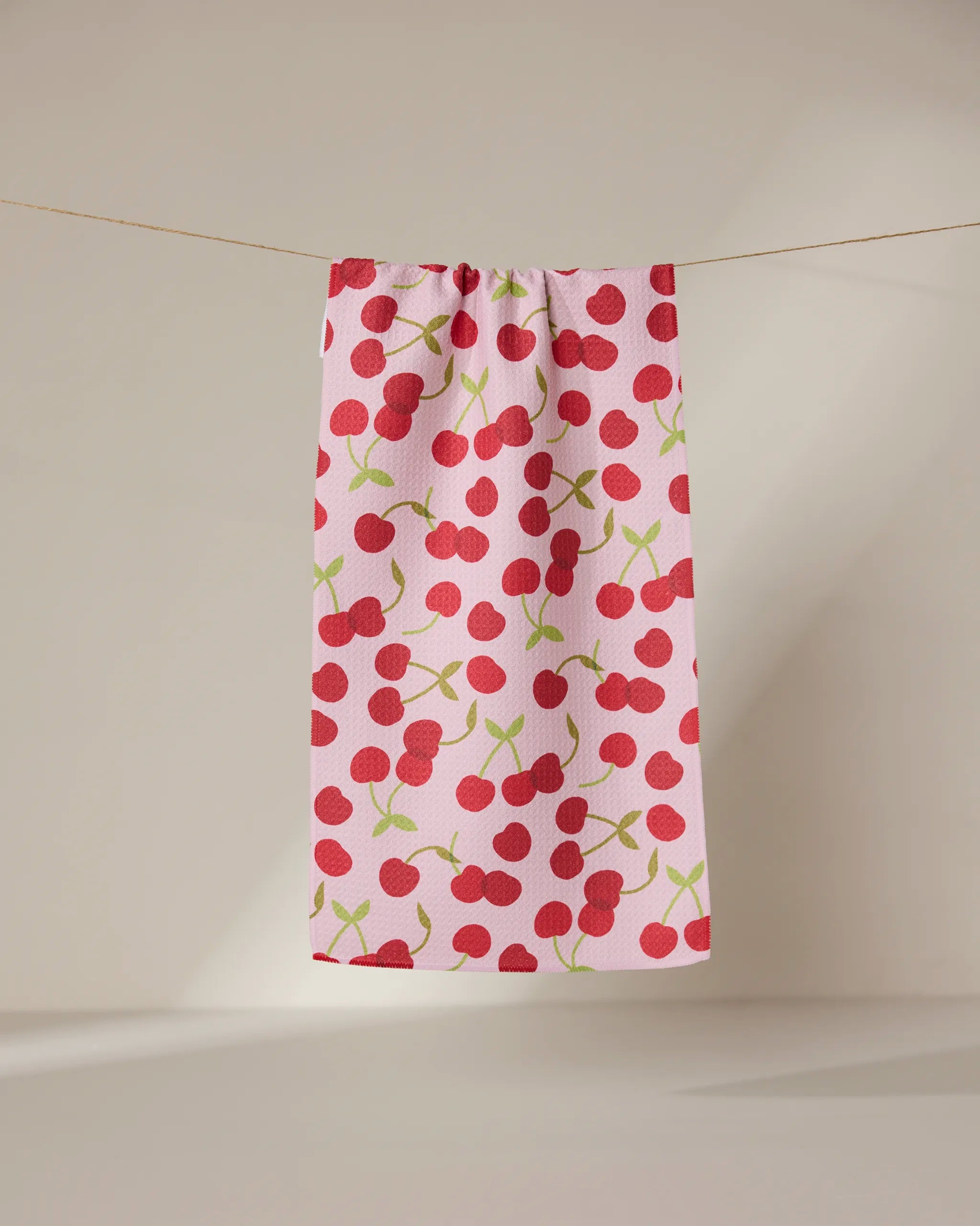 The GEOMETRY Cheery Cherries towel, ultra-absorbent and pink with red cherry and green stem patterns, hangs on a clothesline against a neutral background.
