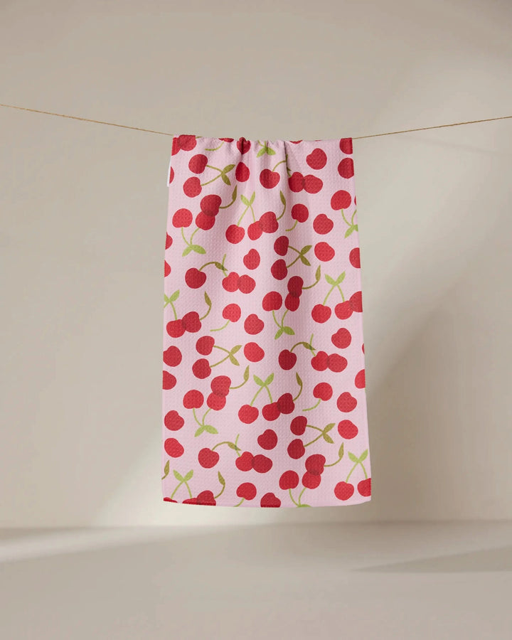 The GEOMETRY Cheery Cherries towel, ultra-absorbent and pink with red cherry and green stem patterns, hangs on a clothesline against a neutral background.