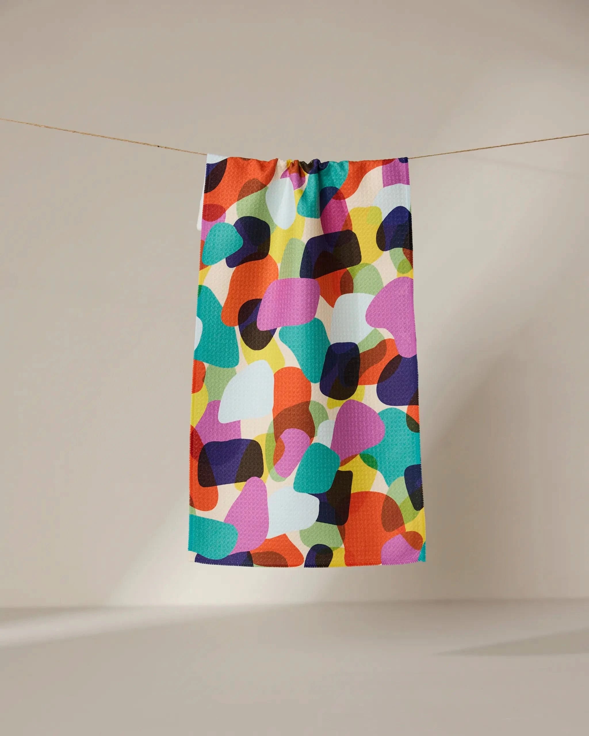 The GEOMETRY Colorful Pebbles tea towel, featuring an ultra-absorbent design and abstract overlapping shapes in vibrant hues, hangs on a line against a plain beige background.
