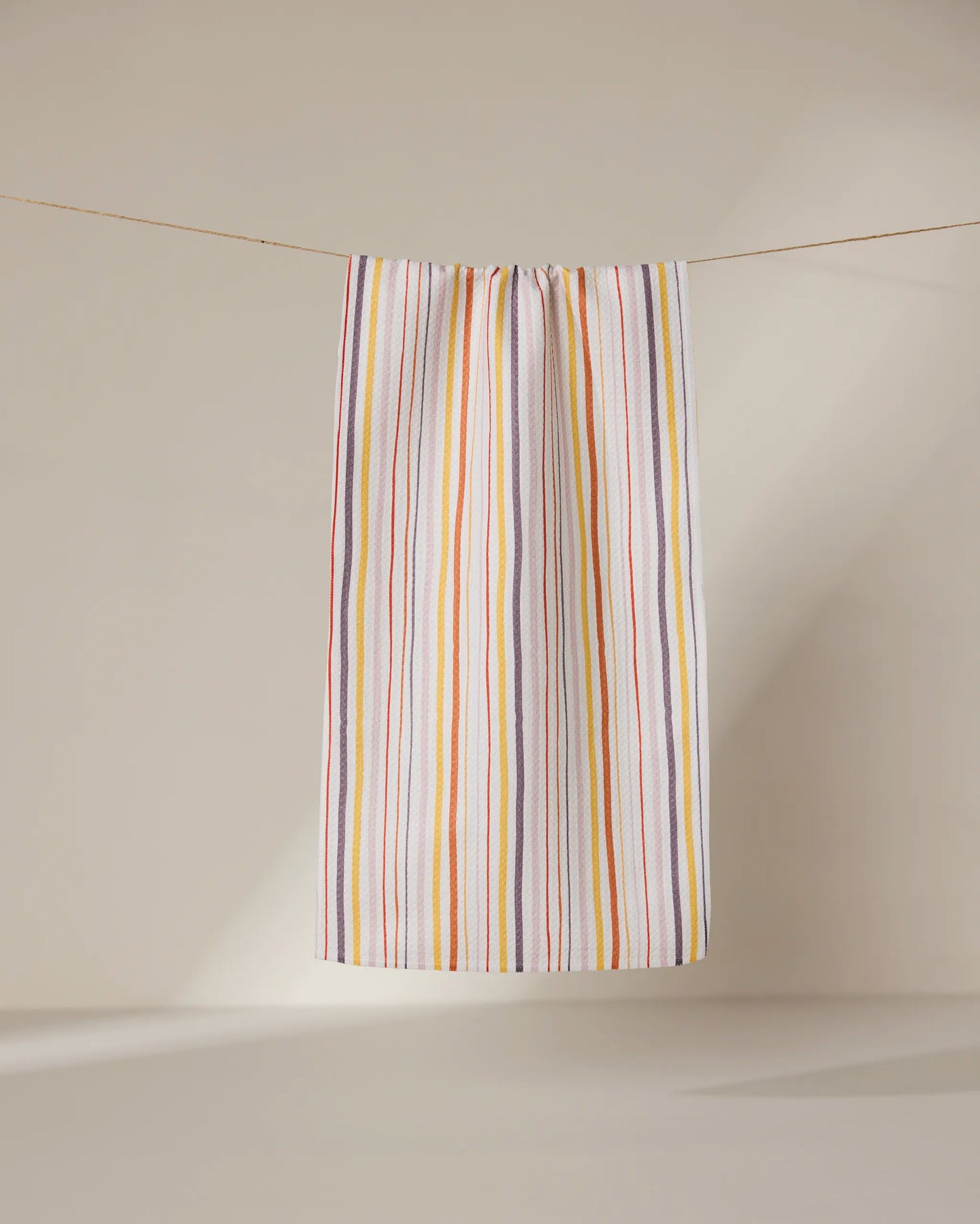 The GEOMETRY Stripe Season towel, ultra-absorbent and white with thin vertical orange, yellow, pink, and purple stripes, hangs from a clothesline against a neutral background.
