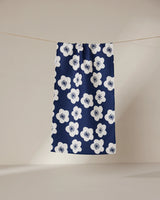 The GEOMETRY Every Sunday kitchen towel, featuring a navy blue color with large white floral patterns, hangs on a clothesline against a plain beige background.