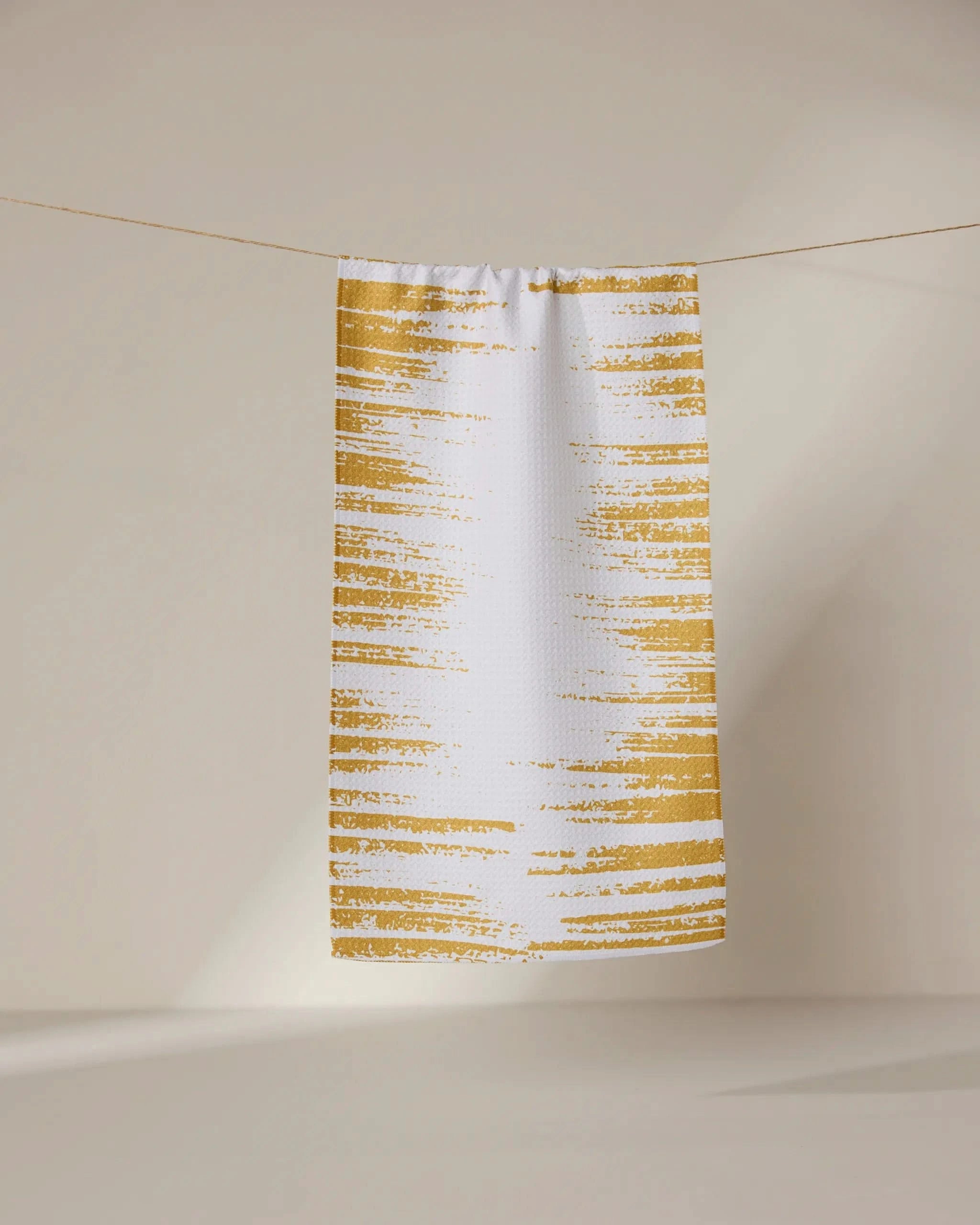 The GEOMETRY Worn Gold towel, ultra-absorbent and white with golden-yellow brushstroke stripes, hangs on a thin line against a softly lit neutral background.