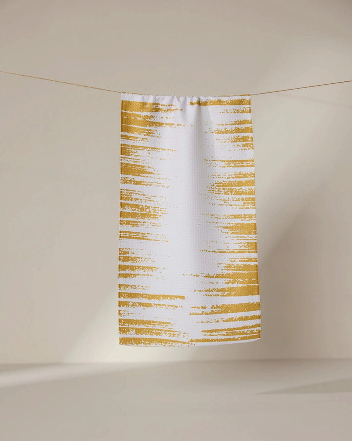 The GEOMETRY Worn Gold towel, ultra-absorbent and white with golden-yellow brushstroke stripes, hangs on a thin line against a softly lit neutral background.