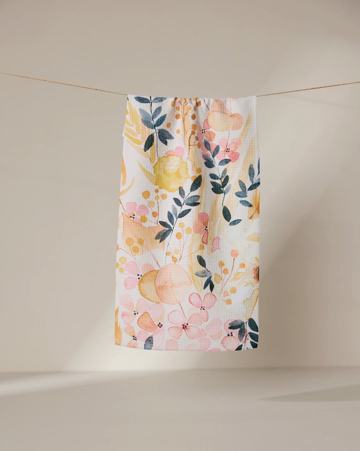 The Sleepy Fields towel by GEOMETRY, featuring a vibrant floral and leaf design in yellow, pink, and dark blue, hangs on a line against a neutral background.
